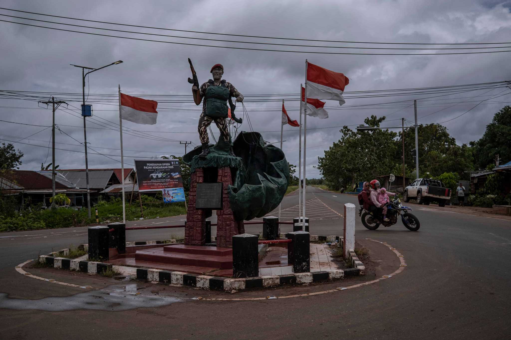 Three people on a moped circle around a roundabout featuring a statue of a soldier